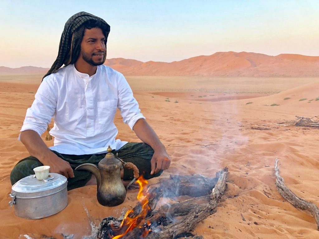 Beduin with headscarf wrapped around his head sitting cross-legged in the sand and looking into the vastness of the desert. In front of him some burning wood and an Omani coffeepot.